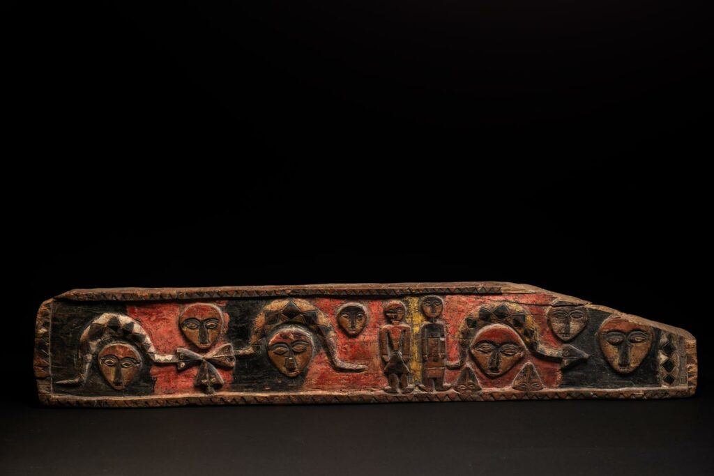 19th-century door lintel from the Paiwan tribe, carved with human heads topped by sacred snakes, along with two standing figures, likely a couple. This Paiwan lintel is painted in black, red, and yellow.
