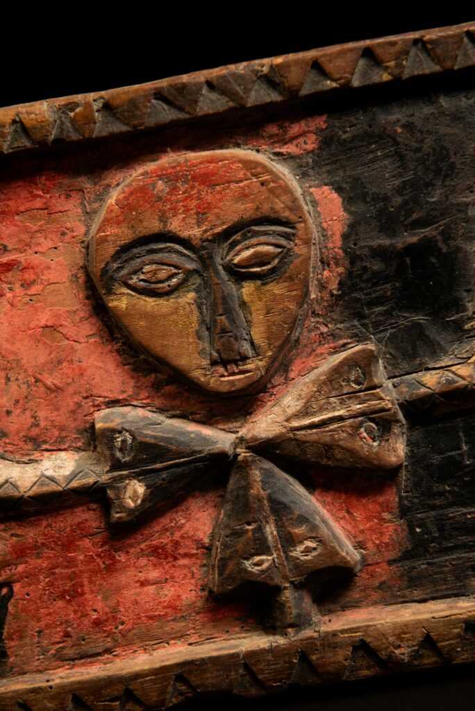 19th-century door lintel from the Paiwan tribe, carved with human heads topped by sacred snakes, along with two standing figures, likely a couple. This Paiwan lintel is painted in black, red, and yellow.