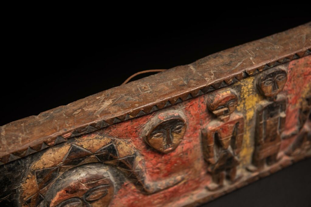 19th-century door lintel from the Paiwan tribe, carved with human heads topped by sacred snakes, along with two standing figures, likely a couple. This Paiwan lintel is painted in black, red, and yellow.