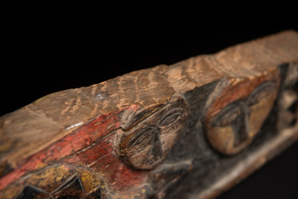 19th-century door lintel from the Paiwan tribe, carved with human heads topped by sacred snakes, along with two standing figures, likely a couple. This Paiwan lintel is painted in black, red, and yellow.