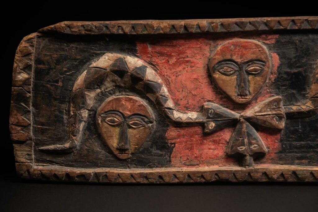19th-century door lintel from the Paiwan tribe, carved with human heads topped by sacred snakes, along with two standing figures, likely a couple. This Paiwan lintel is painted in black, red, and yellow.