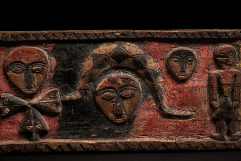 19th-century door lintel from the Paiwan tribe, carved with human heads topped by sacred snakes, along with two standing figures, likely a couple. This Paiwan lintel is painted in black, red, and yellow.