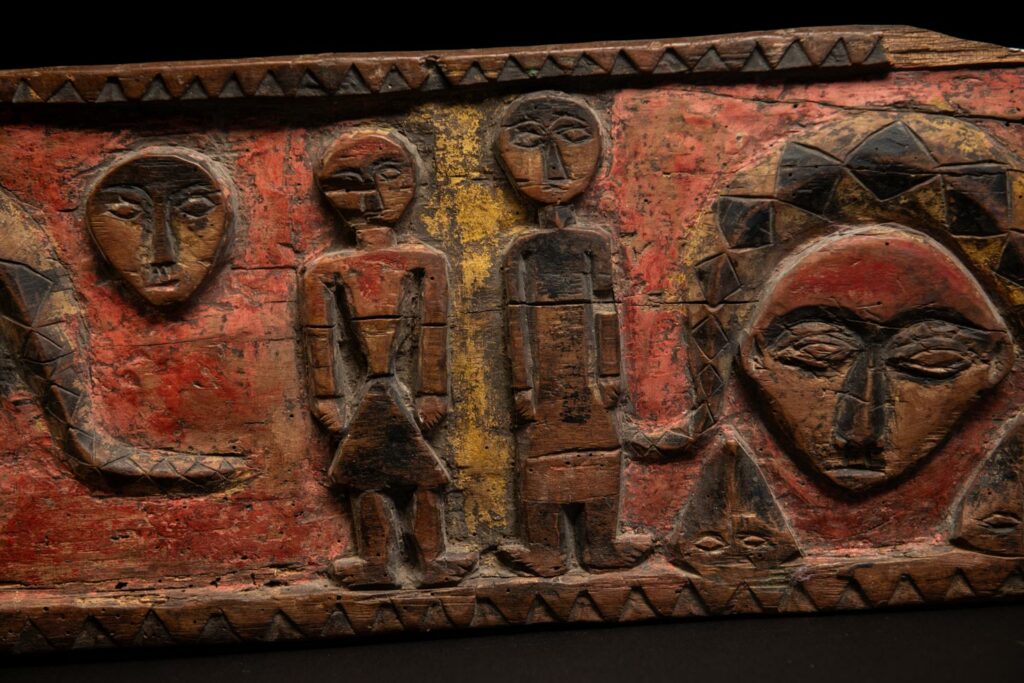 19th-century door lintel from the Paiwan tribe, carved with human heads topped by sacred snakes, along with two standing figures, likely a couple. This Paiwan lintel is painted in black, red, and yellow.