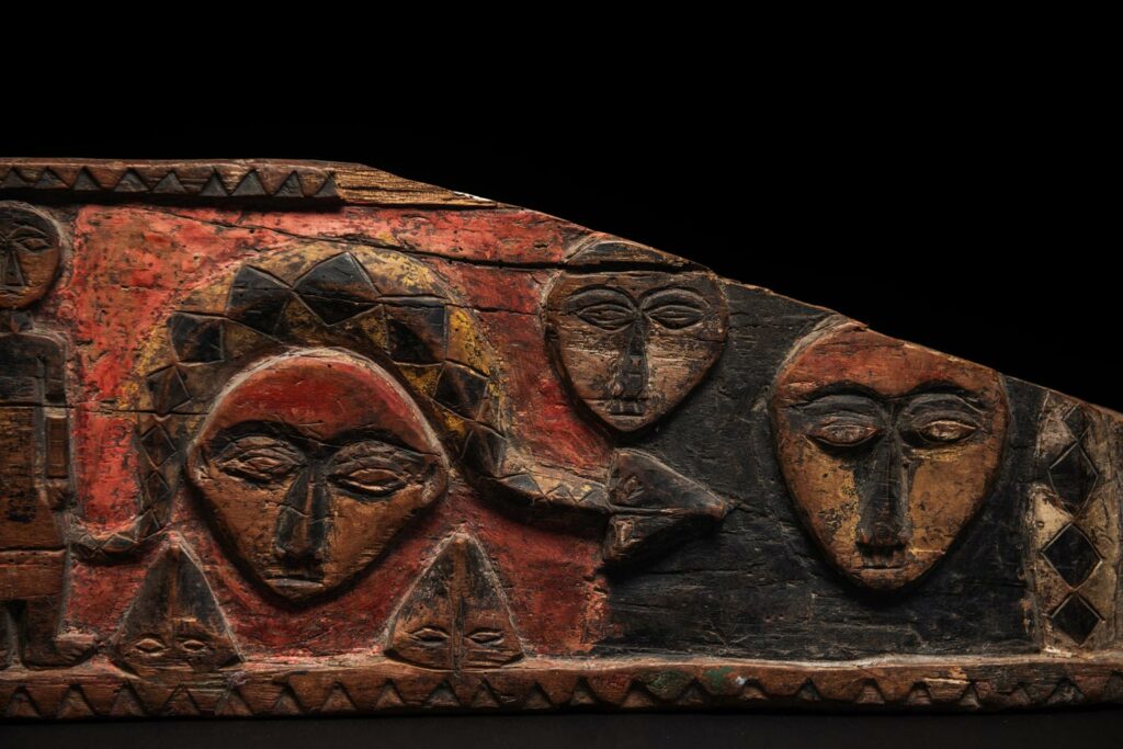 19th-century door lintel from the Paiwan tribe, carved with human heads topped by sacred snakes, along with two standing figures, likely a couple. This Paiwan lintel is painted in black, red, and yellow.