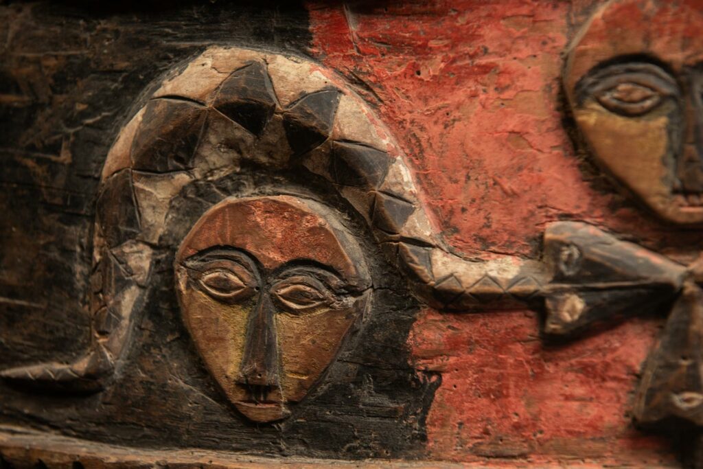19th-century door lintel from the Paiwan tribe, carved with human heads topped by sacred snakes, along with two standing figures, likely a couple. This Paiwan lintel is painted in black, red, and yellow.