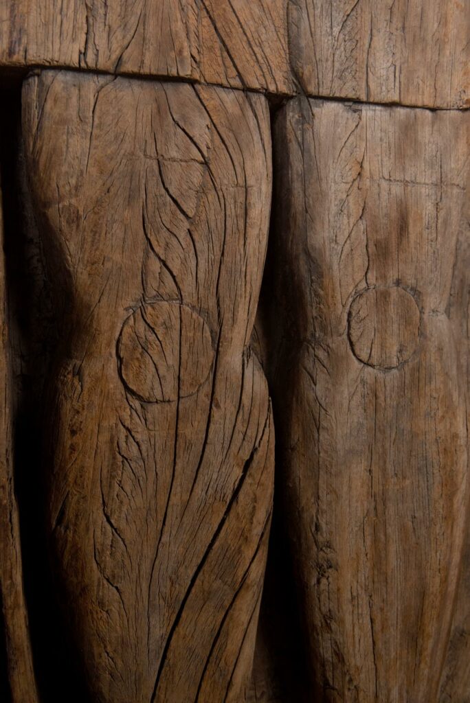 Paiwan tribe: massive 19th-century ancestral panel measuring over 2.5 meters, depicting a hunter wearing a Paiwan-style hat and holding a knife and a spear at his side. The carvings on his torso suggest he is dressed in a ceremonial vest and the shoulder belt traditionally worn by nobles. Due to its size, this panel was likely the central piece of the house.