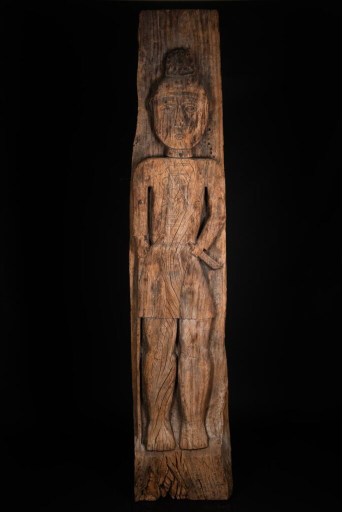 Paiwan tribe: massive 19th-century ancestral panel measuring over 2.5 meters, depicting a hunter wearing a Paiwan-style hat and holding a knife and a spear at his side. The carvings on his torso suggest he is dressed in a ceremonial vest and the shoulder belt traditionally worn by nobles. Due to its size, this panel was likely the central piece of the house.