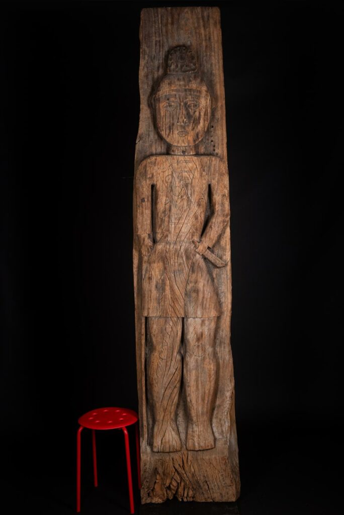 Paiwan tribe: massive 19th-century ancestral panel measuring over 2.5 meters, depicting a hunter wearing a Paiwan-style hat and holding a knife and a spear at his side. The carvings on his torso suggest he is dressed in a ceremonial vest and the shoulder belt traditionally worn by nobles. Due to its size, this panel was likely the central piece of the house.