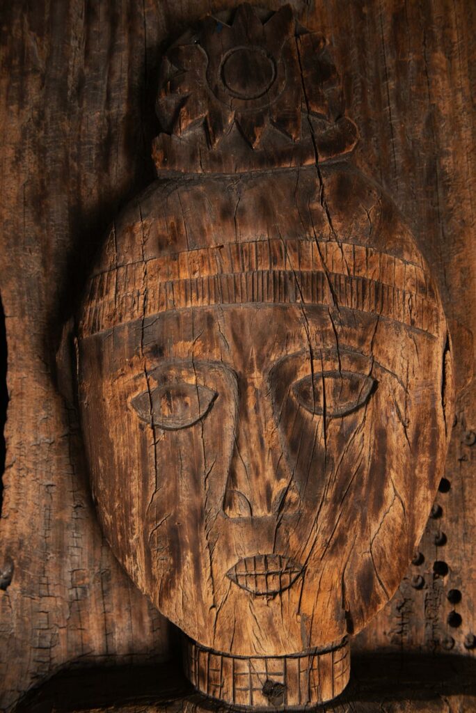 Paiwan tribe: massive 19th-century ancestral panel measuring over 2.5 meters, depicting a hunter wearing a Paiwan-style hat and holding a knife and a spear at his side. The carvings on his torso suggest he is dressed in a ceremonial vest and the shoulder belt traditionally worn by nobles. Due to its size, this panel was likely the central piece of the house.