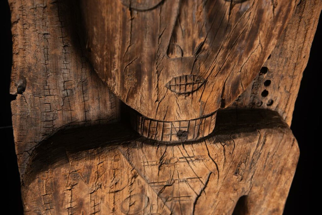 Paiwan tribe: massive 19th-century ancestral panel measuring over 2.5 meters, depicting a hunter wearing a Paiwan-style hat and holding a knife and a spear at his side. The carvings on his torso suggest he is dressed in a ceremonial vest and the shoulder belt traditionally worn by nobles. Due to its size, this panel was likely the central piece of the house.