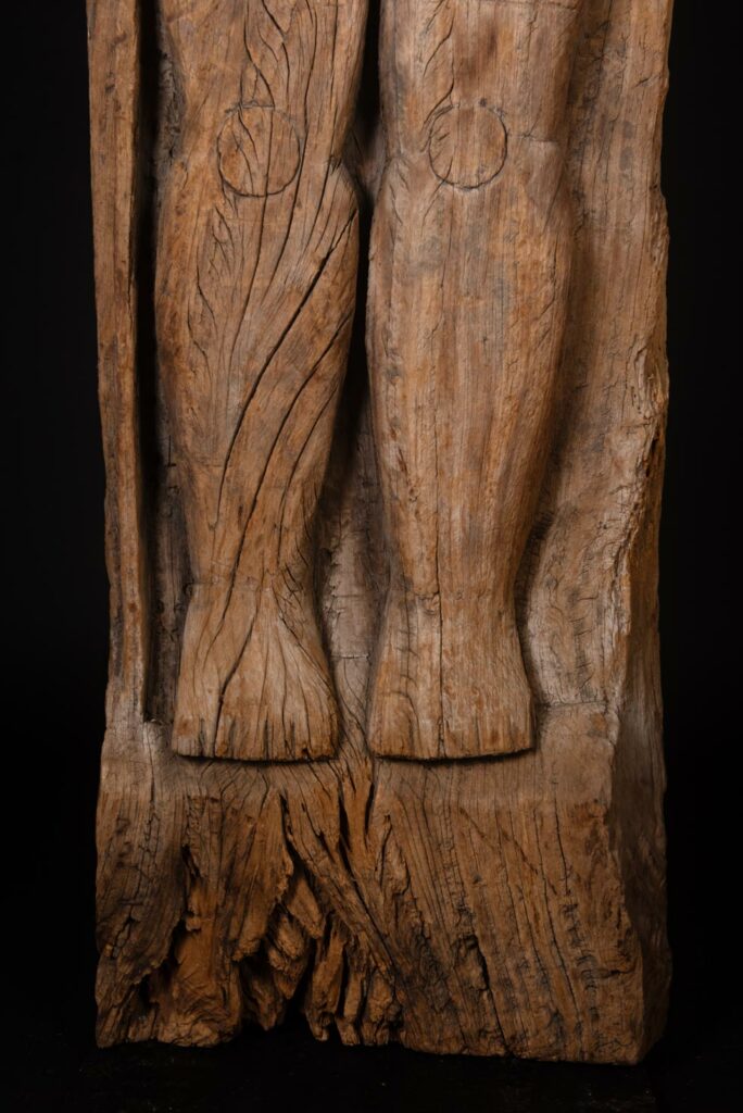 Paiwan tribe: massive 19th-century ancestral panel measuring over 2.5 meters, depicting a hunter wearing a Paiwan-style hat and holding a knife and a spear at his side. The carvings on his torso suggest he is dressed in a ceremonial vest and the shoulder belt traditionally worn by nobles. Due to its size, this panel was likely the central piece of the house.