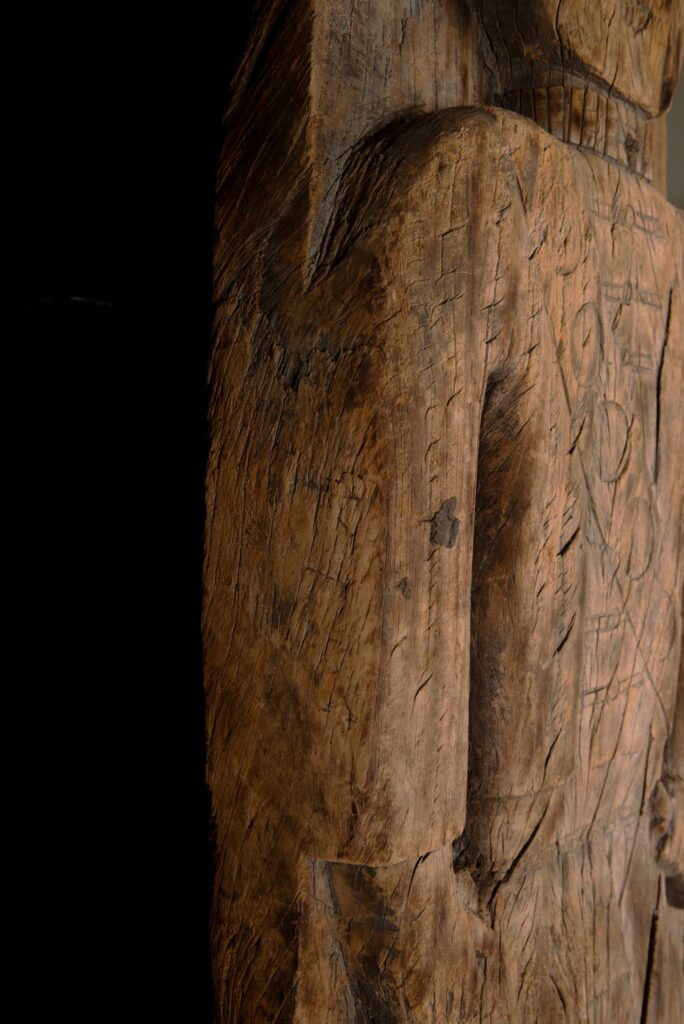 Paiwan tribe: massive 19th-century ancestral panel measuring over 2.5 meters, depicting a hunter wearing a Paiwan-style hat and holding a knife and a spear at his side. The carvings on his torso suggest he is dressed in a ceremonial vest and the shoulder belt traditionally worn by nobles. Due to its size, this panel was likely the central piece of the house.