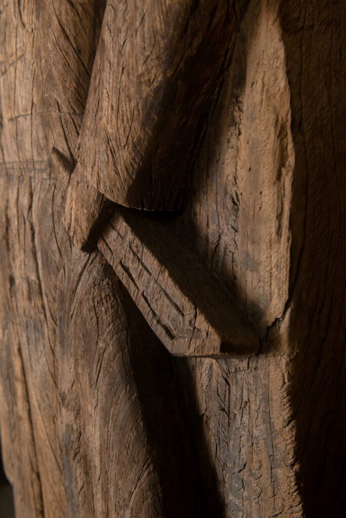 Paiwan tribe: massive 19th-century ancestral panel measuring over 2.5 meters, depicting a hunter wearing a Paiwan-style hat and holding a knife and a spear at his side. The carvings on his torso suggest he is dressed in a ceremonial vest and the shoulder belt traditionally worn by nobles. Due to its size, this panel was likely the central piece of the house.