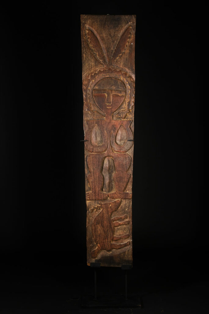 Ancient panel from the Paiwan tribe, carved with a stylized ancestor wearing a two-headed snake headdress — a symbol of noble lineage — and eagle feathers, representing strength in battle. A deer lies at his feet.