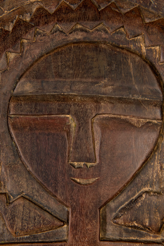 Ancient panel from the Paiwan tribe, carved with a stylized ancestor wearing a two-headed snake headdress — a symbol of noble lineage — and eagle feathers, representing strength in battle. A deer lies at his feet.