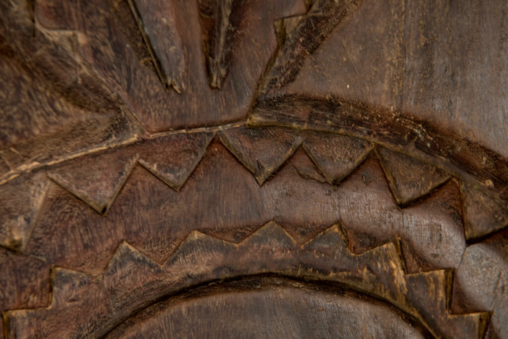 Ancient panel from the Paiwan tribe, carved with a stylized ancestor wearing a two-headed snake headdress — a symbol of noble lineage — and eagle feathers, representing strength in battle. A deer lies at his feet.