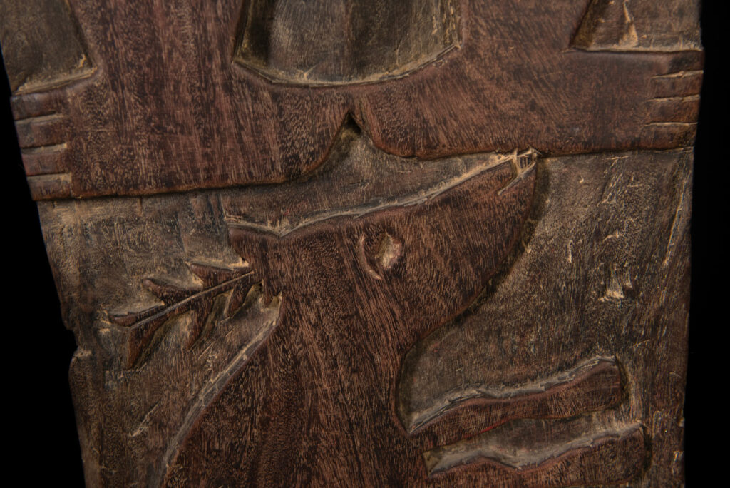 Ancient panel from the Paiwan tribe, carved with a stylized ancestor wearing a two-headed snake headdress — a symbol of noble lineage — and eagle feathers, representing strength in battle. A deer lies at his feet.