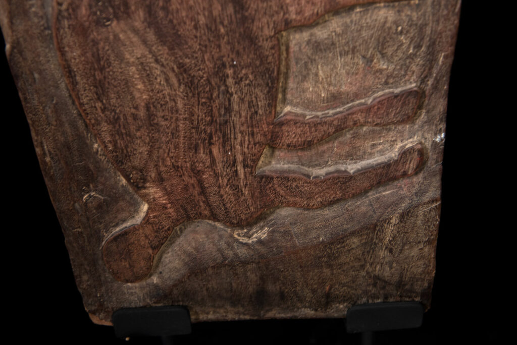 Ancient panel from the Paiwan tribe, carved with a stylized ancestor wearing a two-headed snake headdress — a symbol of noble lineage — and eagle feathers, representing strength in battle. A deer lies at his feet.