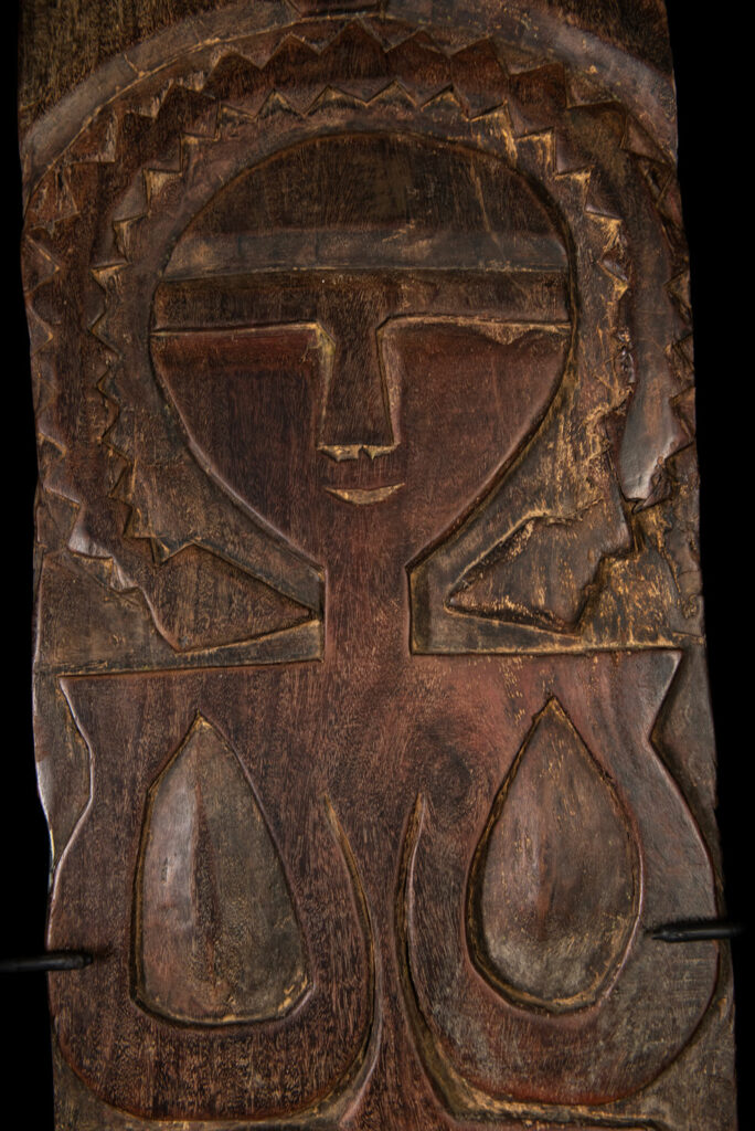 Ancient panel from the Paiwan tribe, carved with a stylized ancestor wearing a two-headed snake headdress — a symbol of noble lineage — and eagle feathers, representing strength in battle. A deer lies at his feet.