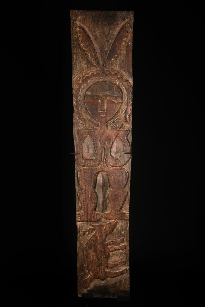 Ancient panel from the Paiwan tribe, carved with a stylized ancestor wearing a two-headed snake headdress — a symbol of noble lineage — and eagle feathers, representing strength in battle. A deer lies at his feet.