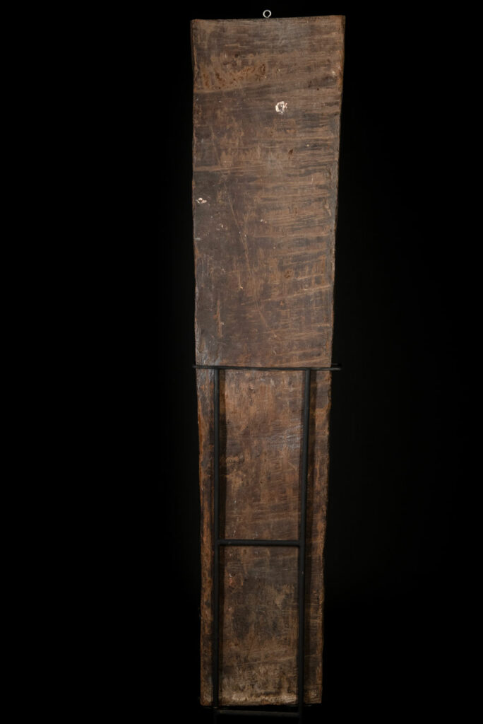 Ancient panel from the Paiwan tribe, carved with a stylized ancestor wearing a two-headed snake headdress — a symbol of noble lineage — and eagle feathers, representing strength in battle. A deer lies at his feet.