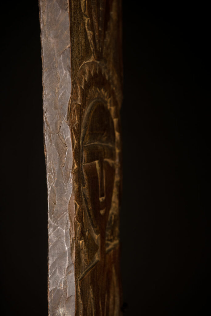 Ancient panel from the Paiwan tribe, carved with a stylized ancestor wearing a two-headed snake headdress — a symbol of noble lineage — and eagle feathers, representing strength in battle. A deer lies at his feet.