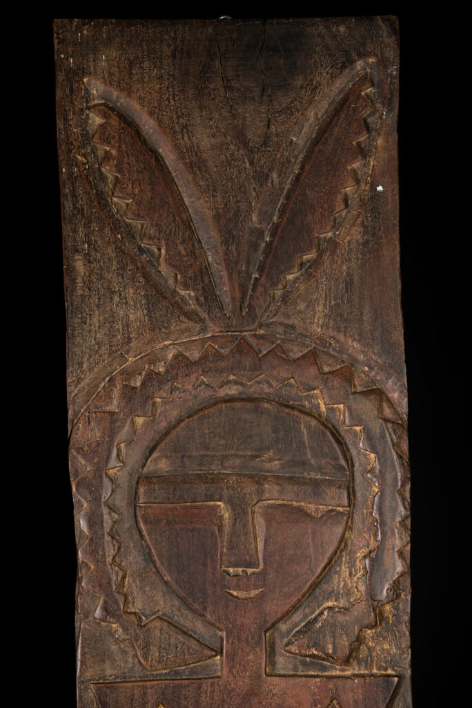 Ancient panel from the Paiwan tribe, carved with a stylized ancestor wearing a two-headed snake headdress — a symbol of noble lineage — and eagle feathers, representing strength in battle. A deer lies at his feet.