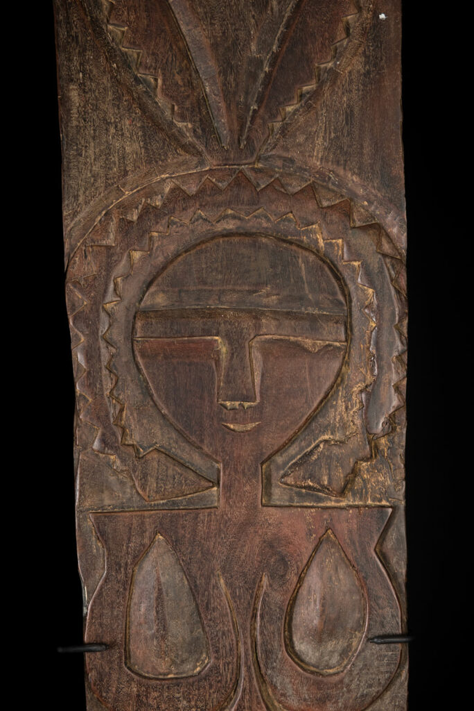 Ancient panel from the Paiwan tribe, carved with a stylized ancestor wearing a two-headed snake headdress — a symbol of noble lineage — and eagle feathers, representing strength in battle. A deer lies at his feet.