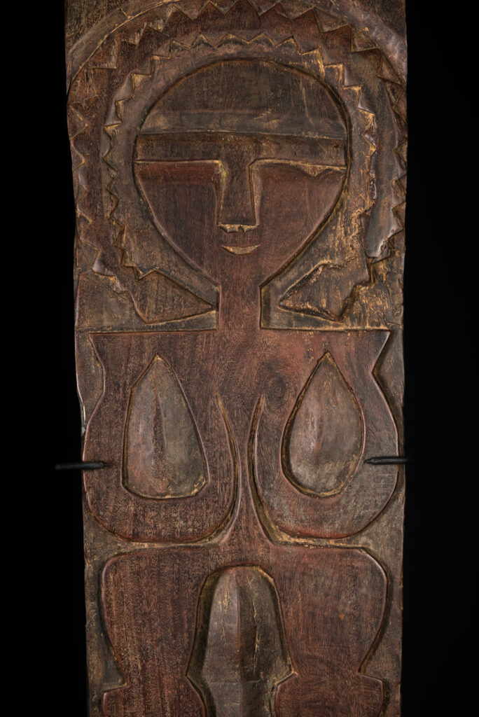 Ancient panel from the Paiwan tribe, carved with a stylized ancestor wearing a two-headed snake headdress — a symbol of noble lineage — and eagle feathers, representing strength in battle. A deer lies at his feet.