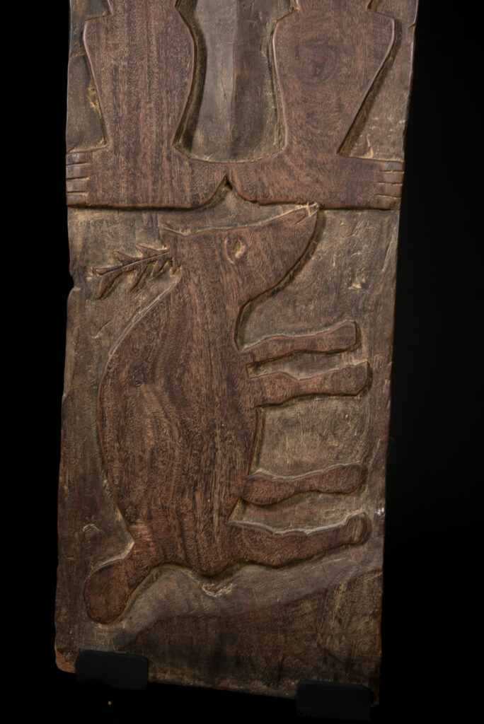 Ancient panel from the Paiwan tribe, carved with a stylized ancestor wearing a two-headed snake headdress — a symbol of noble lineage — and eagle feathers, representing strength in battle. A deer lies at his feet.