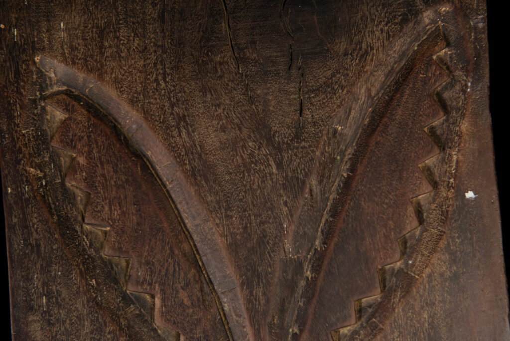 Ancient panel from the Paiwan tribe, carved with a stylized ancestor wearing a two-headed snake headdress — a symbol of noble lineage — and eagle feathers, representing strength in battle. A deer lies at his feet.