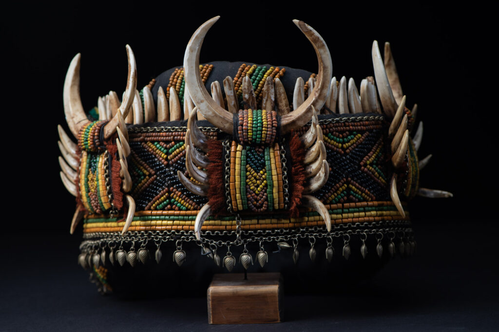 Women's headdress from the Paiwan tribe, made of green, yellow, red, and black beads arranged in geometric patterns, surrounded by chains and topped with boar tusks.