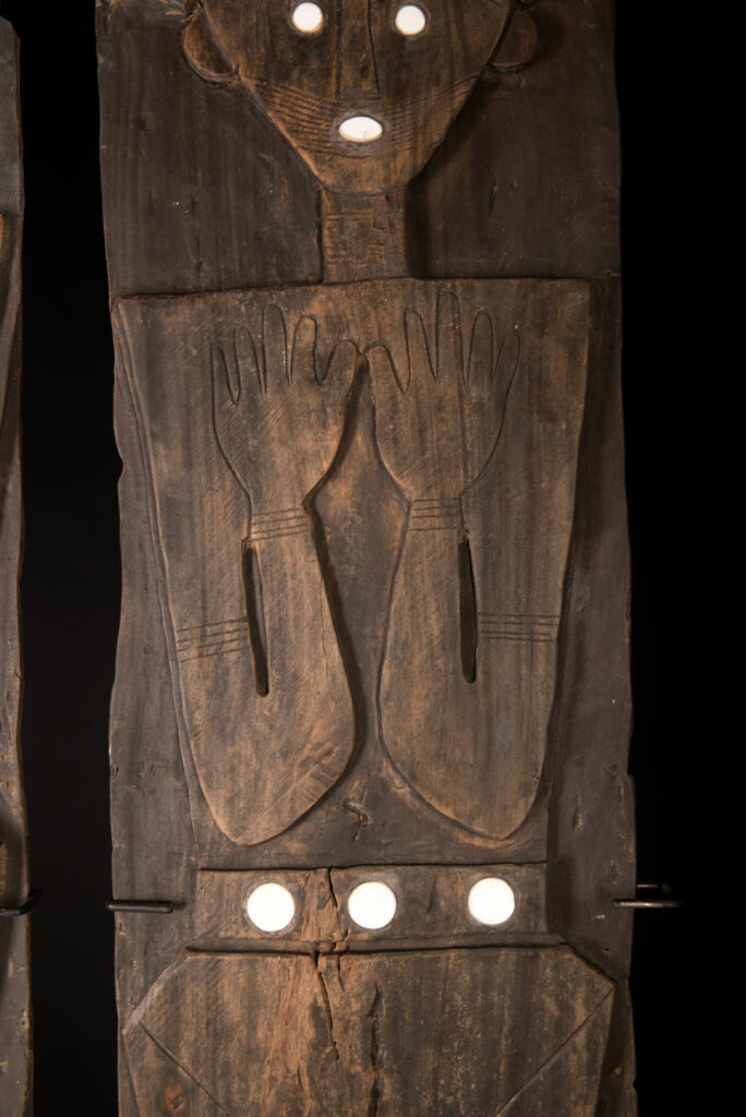 Pair of ancestor panels from the Paiwan tribe, depicting a male and a female figure, both wearing feathered headdresses — symbols of valor and vigor. The panels are inlaid with porcelain circles at the eyes, mouth, and waist, representing the ceremonial belts they wear.