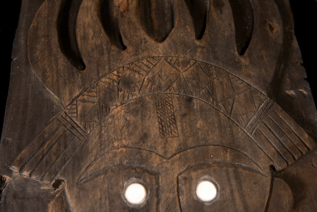 Pair of ancestor panels from the Paiwan tribe, depicting a male and a female figure, both wearing feathered headdresses — symbols of valor and vigor. The panels are inlaid with porcelain circles at the eyes, mouth, and waist, representing the ceremonial belts they wear.