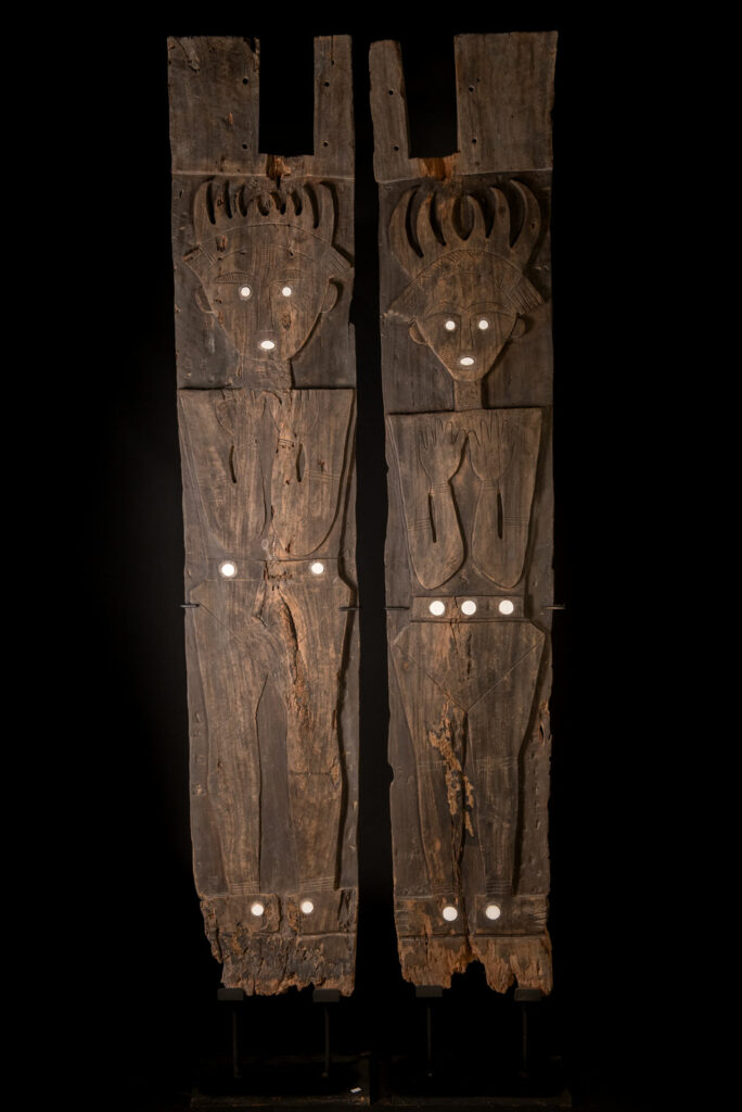 Pair of ancestor panels from the Paiwan tribe, depicting a male and a female figure, both wearing feathered headdresses — symbols of valor and vigor. The panels are inlaid with porcelain circles at the eyes, mouth, and waist, representing the ceremonial belts they wear.