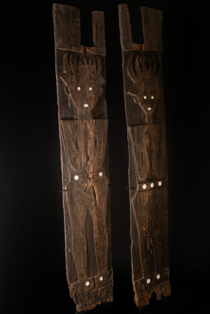 Pair of ancestor panels from the Paiwan tribe, depicting a male and a female figure, both wearing feathered headdresses — symbols of valor and vigor. The panels are inlaid with porcelain circles at the eyes, mouth, and waist, representing the ceremonial belts they wear.
