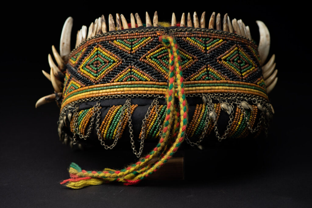 Women's headdress from the Paiwan tribe, made of green, yellow, red, and black beads arranged in geometric patterns, surrounded by chains and topped with boar tusks.