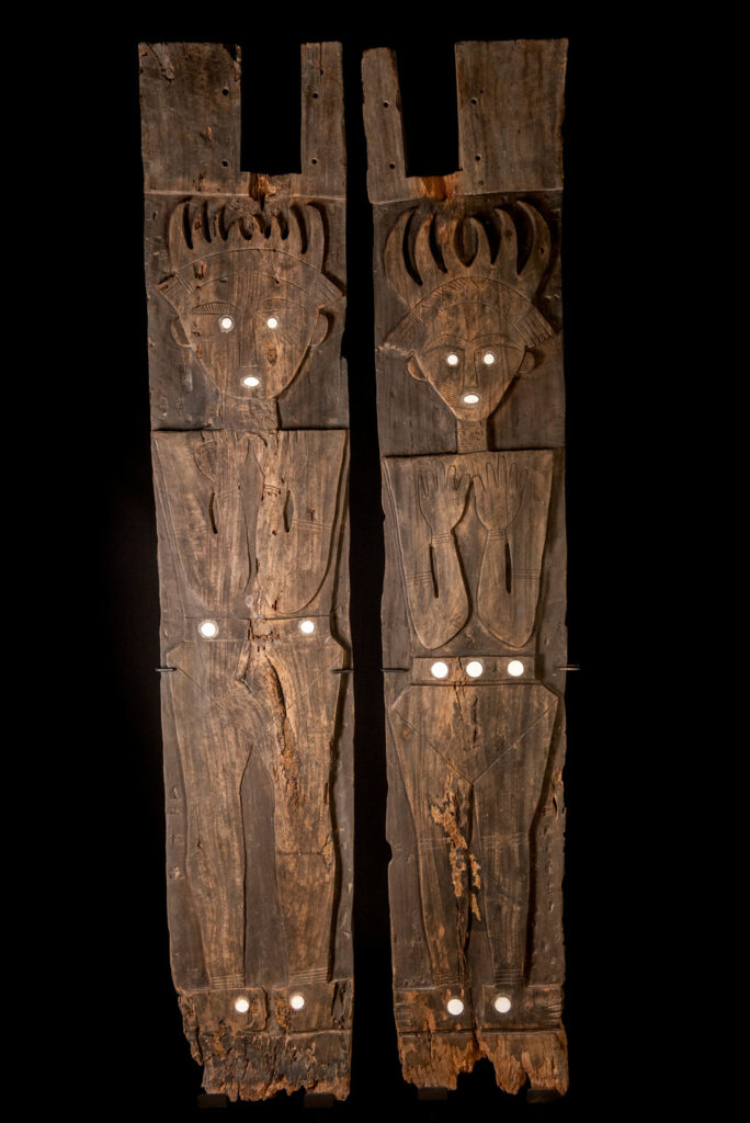 Pair of ancestor panels from the Paiwan tribe, depicting a male and a female figure, both wearing feathered headdresses — symbols of valor and vigor. The panels are inlaid with porcelain circles at the eyes, mouth, and waist, representing the ceremonial belts they wear.