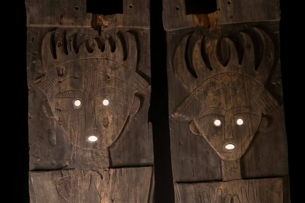 Pair of ancestor panels from the Paiwan tribe, depicting a male and a female figure, both wearing feathered headdresses — symbols of valor and vigor. The panels are inlaid with porcelain circles at the eyes, mouth, and waist, representing the ceremonial belts they wear.