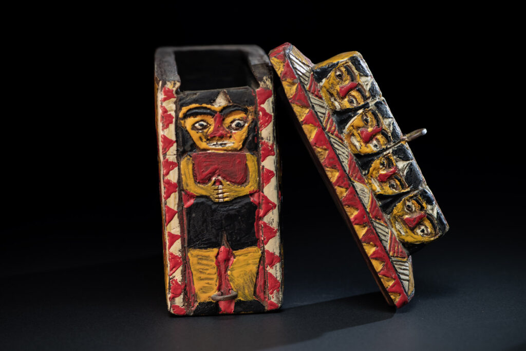 Paiwan tribe shaman box features a sliding lid, carved with human heads and painted in red, yellow, white, and black. Standing figure is carved on the other sides.