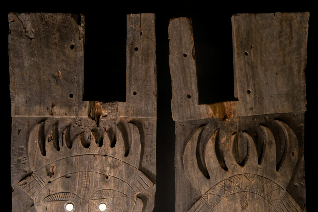 Pair of ancestor panels from the Paiwan tribe, depicting a male and a female figure, both wearing feathered headdresses — symbols of valor and vigor. The panels are inlaid with porcelain circles at the eyes, mouth, and waist, representing the ceremonial belts they wear.