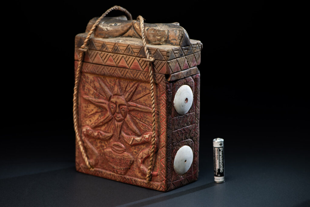 Ancient shaman box from the Paiwan tribe, stained red, with a sliding lid carved with snakes face. One side is carved with two tattooed hands, while the other features a sun, a pot, and two snakes — depicting the myth of the Paiwan people's origin from the union of a pot and a snake. The sides are inlaid with circular shell ornaments.