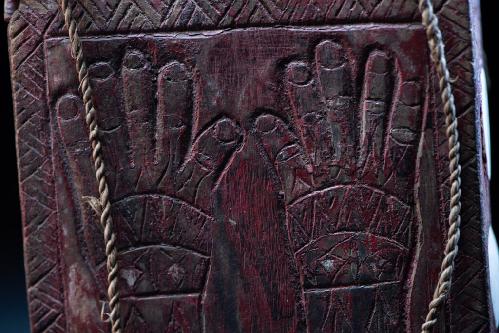 Ancient shaman box from the Paiwan tribe, stained red, with a sliding lid carved with snakes face. One side is carved with two tattooed hands, while the other features a sun, a pot, and two snakes — depicting the myth of the Paiwan people's origin from the union of a pot and a snake. The sides are inlaid with circular shell ornaments.