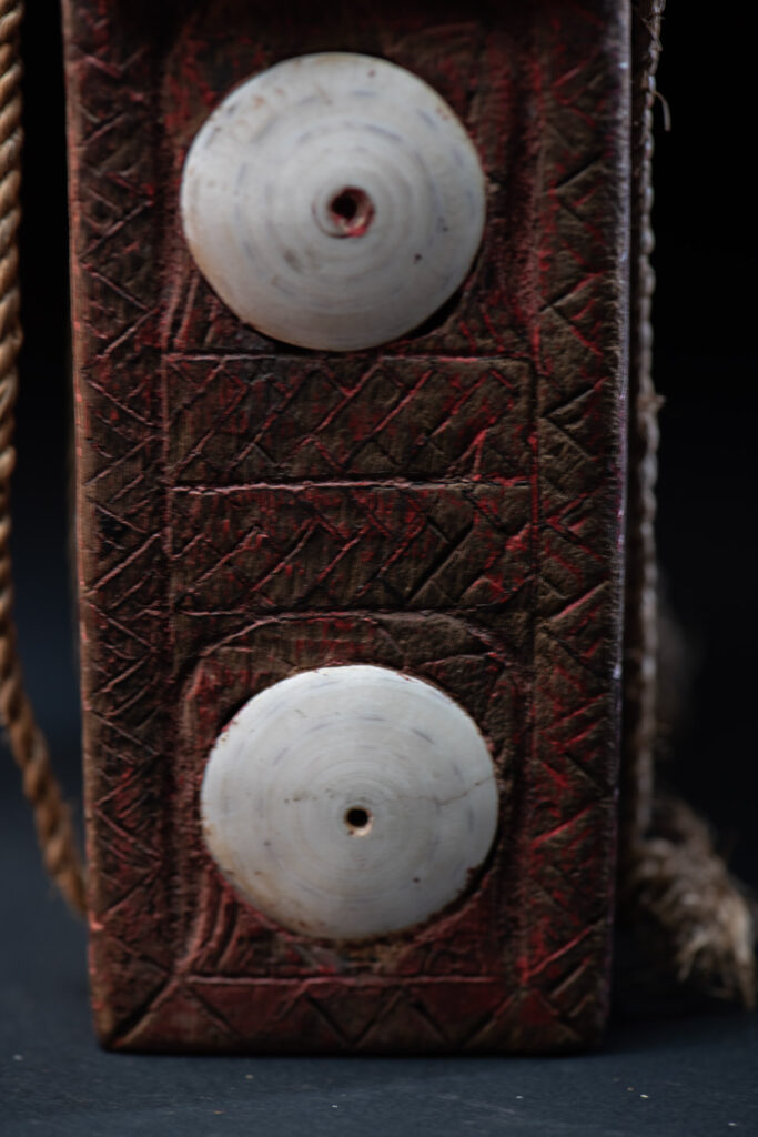 Ancient shaman box from the Paiwan tribe, stained red, with a sliding lid carved with snakes face. One side is carved with two tattooed hands, while the other features a sun, a pot, and two snakes — depicting the myth of the Paiwan people's origin from the union of a pot and a snake. The sides are inlaid with circular shell ornaments.