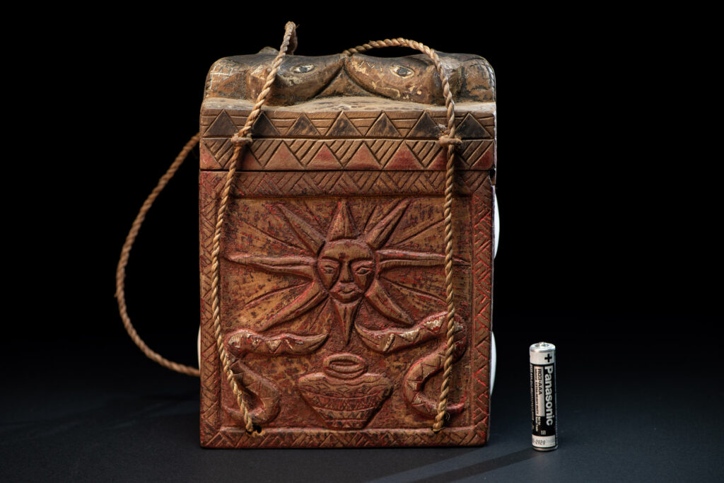 Ancient shaman box from the Paiwan tribe, stained red, with a sliding lid carved with snakes face. One side is carved with two tattooed hands, while the other features a sun, a pot, and two snakes — depicting the myth of the Paiwan people's origin from the union of a pot and a snake. The sides are inlaid with circular shell ornaments.