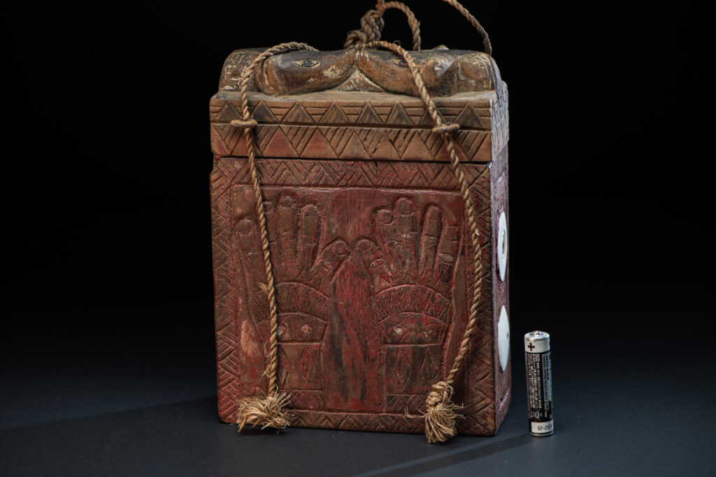 Ancient shaman box from the Paiwan tribe, stained red, with a sliding lid carved with snakes face. One side is carved with two tattooed hands, while the other features a sun, a pot, and two snakes — depicting the myth of the Paiwan people's origin from the union of a pot and a snake. The sides are inlaid with circular shell ornaments.