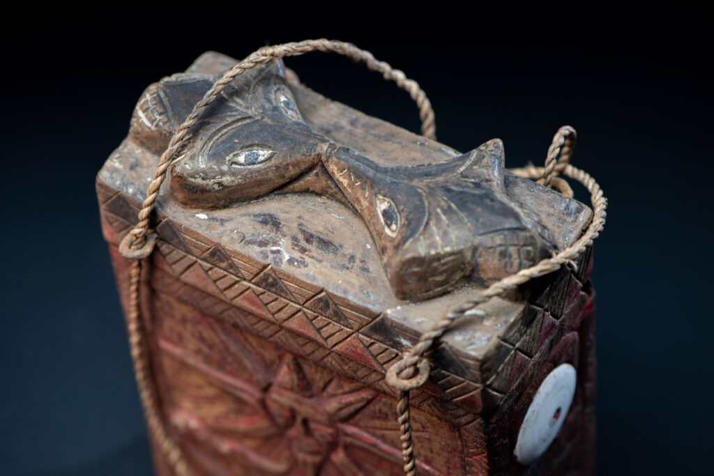 Ancient shaman box from the Paiwan tribe, stained red, with a sliding lid carved with snakes face. One side is carved with two tattooed hands, while the other features a sun, a pot, and two snakes — depicting the myth of the Paiwan people's origin from the union of a pot and a snake. The sides are inlaid with circular shell ornaments.