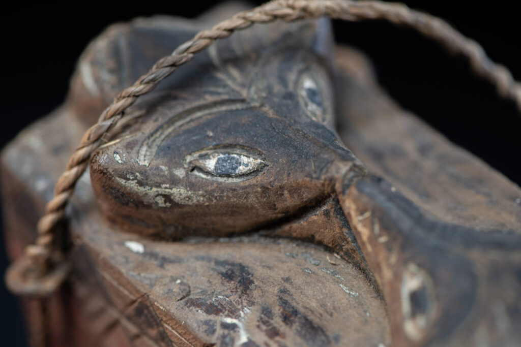 Ancient shaman box from the Paiwan tribe, stained red, with a sliding lid carved with snakes face. One side is carved with two tattooed hands, while the other features a sun, a pot, and two snakes — depicting the myth of the Paiwan people's origin from the union of a pot and a snake. The sides are inlaid with circular shell ornaments.