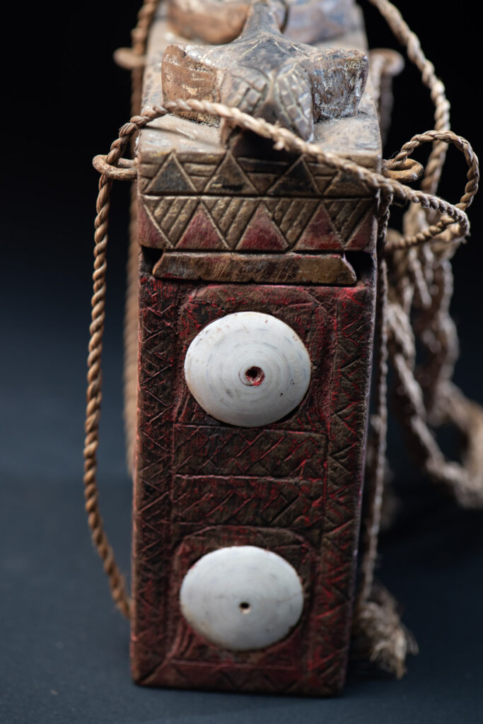 Ancient shaman box from the Paiwan tribe, stained red, with a sliding lid carved with snakes face. One side is carved with two tattooed hands, while the other features a sun, a pot, and two snakes — depicting the myth of the Paiwan people's origin from the union of a pot and a snake. The sides are inlaid with circular shell ornaments.