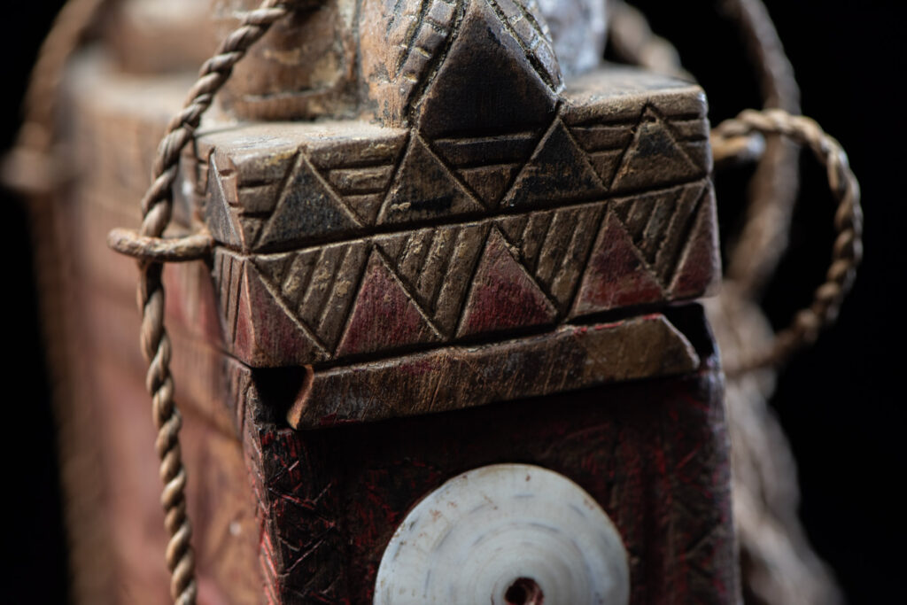 Ancient shaman box from the Paiwan tribe, stained red, with a sliding lid carved with snakes face. One side is carved with two tattooed hands, while the other features a sun, a pot, and two snakes — depicting the myth of the Paiwan people's origin from the union of a pot and a snake. The sides are inlaid with circular shell ornaments.